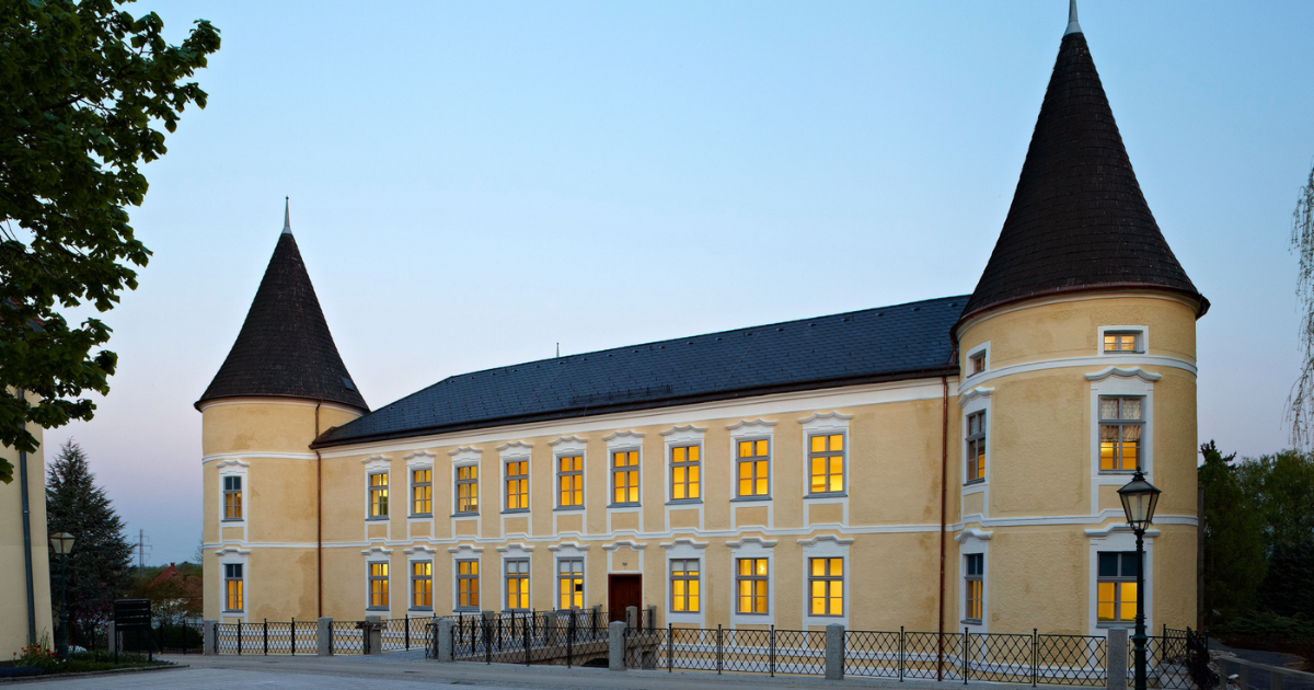 Historische gelbe Schlossanlage mit zwei runden Türmen, Fensterreihen mit warmem Licht, Eingangstor und schmiedeeisernem Zaun.