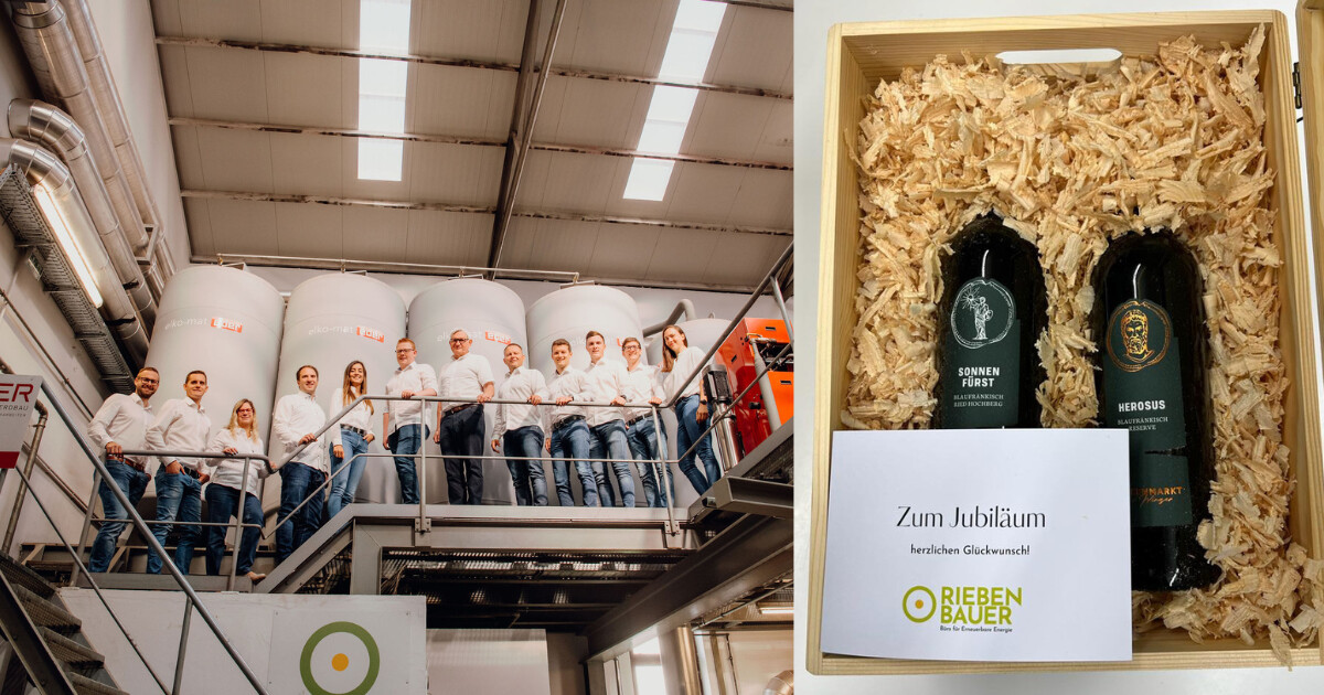 Gruppenbild von Mitarbeitenden in weißen Hemden auf einer Metallbühne in einer Industriehalle; rechts Holzkiste mit zwei Flaschen und Grußkarte.