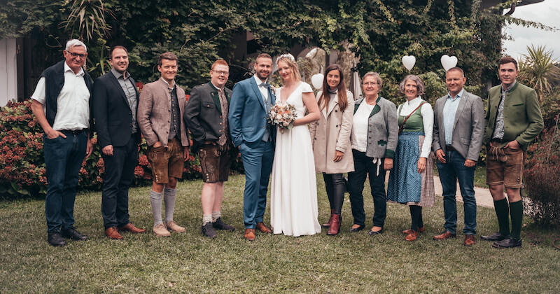 Gruppe im Garten bei einer Hochzeit: eine Person in Weiß, eine Person im blauen Anzug, weitere Gäste in Trachten und formeller Kleidung; Herzballons.