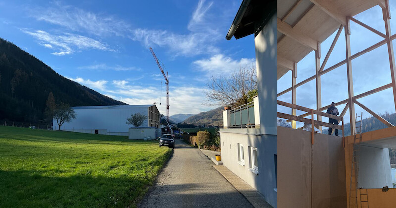 Bauareal in ländlicher Landschaft mit Kran, Rohbau und Holzgerüst; Bauarbeiter auf dem Gerüst, grüne Wiese und Bergkulisse.