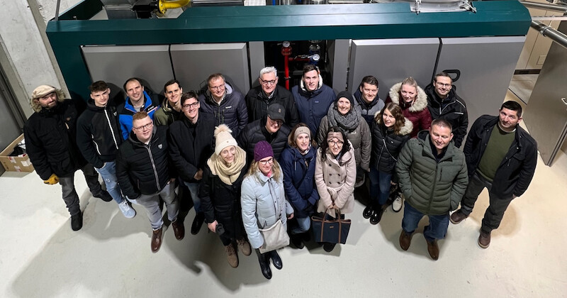 Gruppe von Mitarbeitenden in Winterjacken vor einer großen Industriemaschine, von oben aufgenommen.