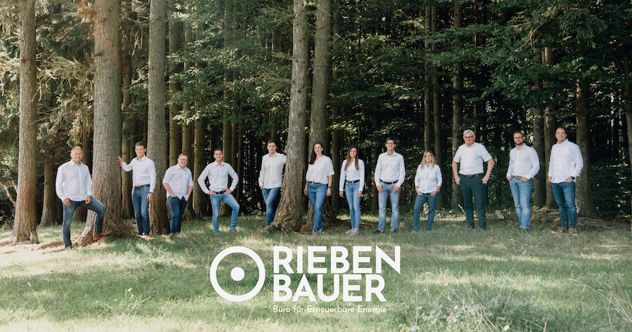Teamfoto des Büros für Erneuerbare Energie RIEBEN BAUER im Wald; Gruppe in Jeans und hellen Oberteilen, Logo unten mittig.