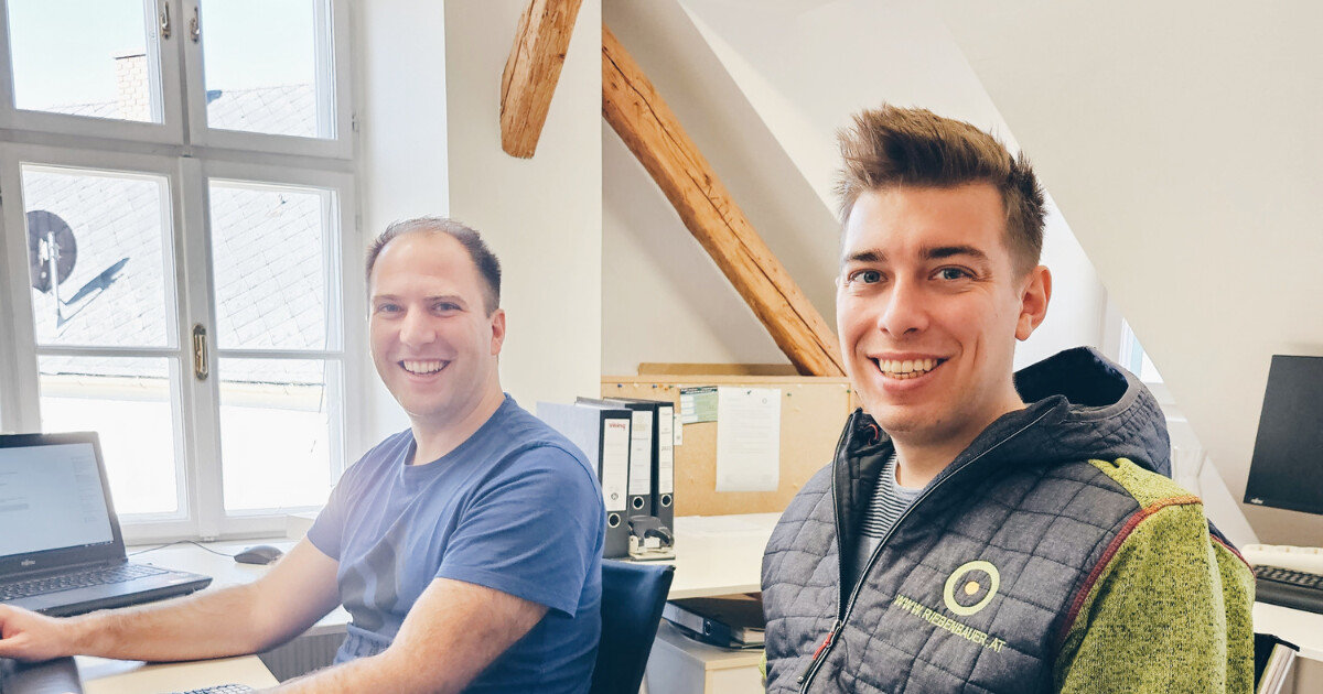 Zwei Personen arbeiten in einem Büro an Computern und lächeln freundlich.