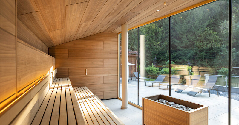 Moderne Sauna aus Holz mit langen Bänken, indirekter Beleuchtung und Glasfront zur Terrasse mit Liegen; Ofen mit Steinen sichtbar.