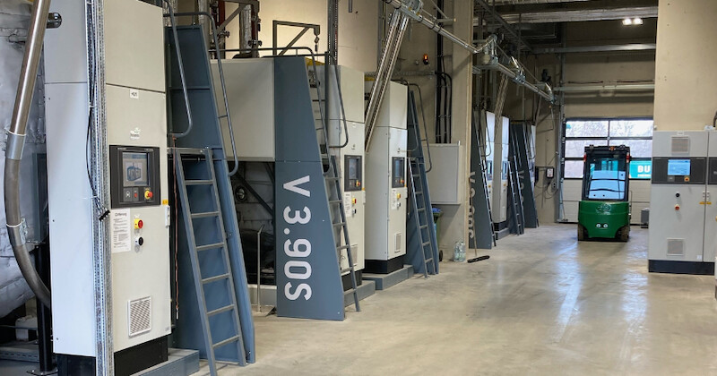 Industriehalle mit Reihen von Maschinen, Leitungen und Metallrahmen; grüner Gabelstapler im Hintergrund.
