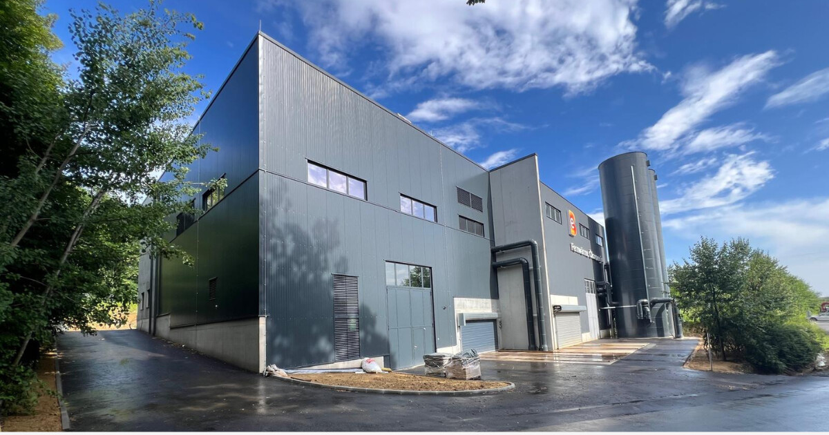Moderne Industrieanlage mit dunkelgrauer Metallfassade, großem Silo, Zufahrt, Bäumen links und blauem Himmel.