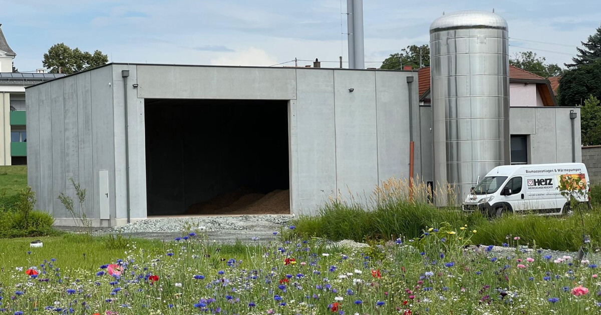 Industrieanlage mit offener Torfront, silbernem Pufferspeicher, weißem Lieferwagen und bunter Blumenwiese im Vordergrund.