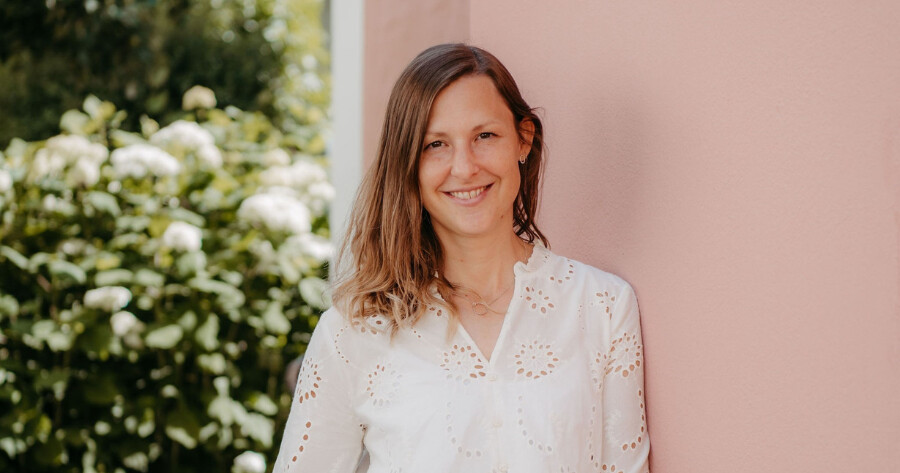 Person mit braunen Haaren in weißer Lochstickerei-Bluse vor rosa Wand, lächelnd; grüner Garten im Hintergrund.