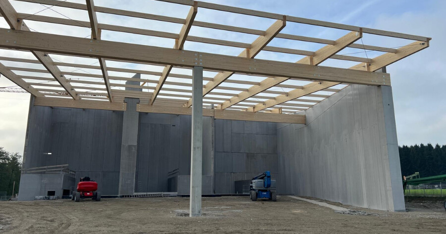 Rohbau einer Halle mit Holzdachrahmen, Betonwänden und Stahlstützen; rote und blaue Baugeräte stehen auf dem Baugrund.