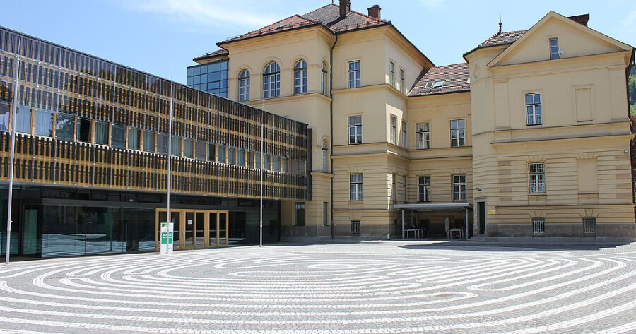 Historische gelbe Gebäudefassade neben moderner Glasfassade; gepflasterter Platz mit konzentrischen Kreismustern vor dem Eingang.