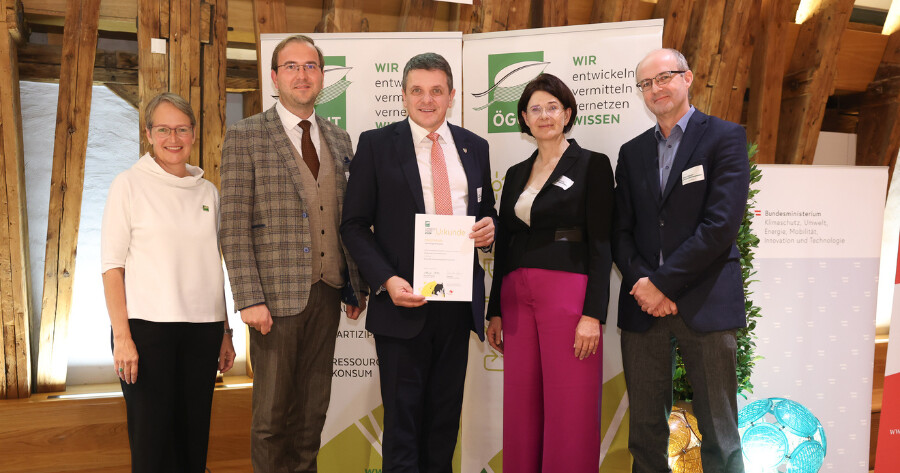 Fünf Personen posieren bei einer Preisverleihung; in der Mitte hält eine Person eine Urkunde, Banner mit Logos im Hintergrund.