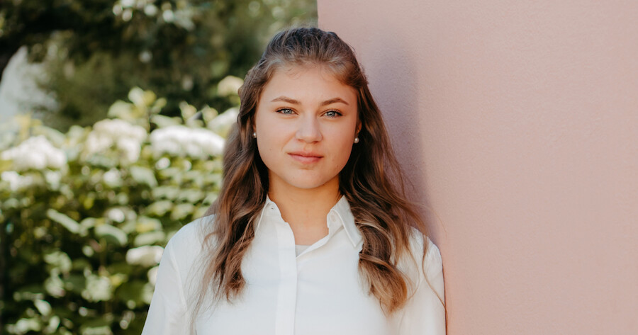 Person mit blonden Haaren und weißem Hemd steht an rosa Wand, grüne Gartenlandschaft im Hintergrund.