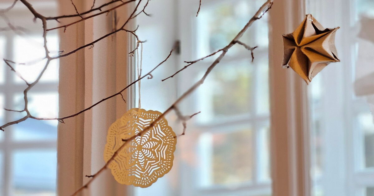 Braune Zweige mit hängenden Papierornamenten: filigranes rundes Mandala und origami-Stern; unscharfer Fensterhintergrund.
