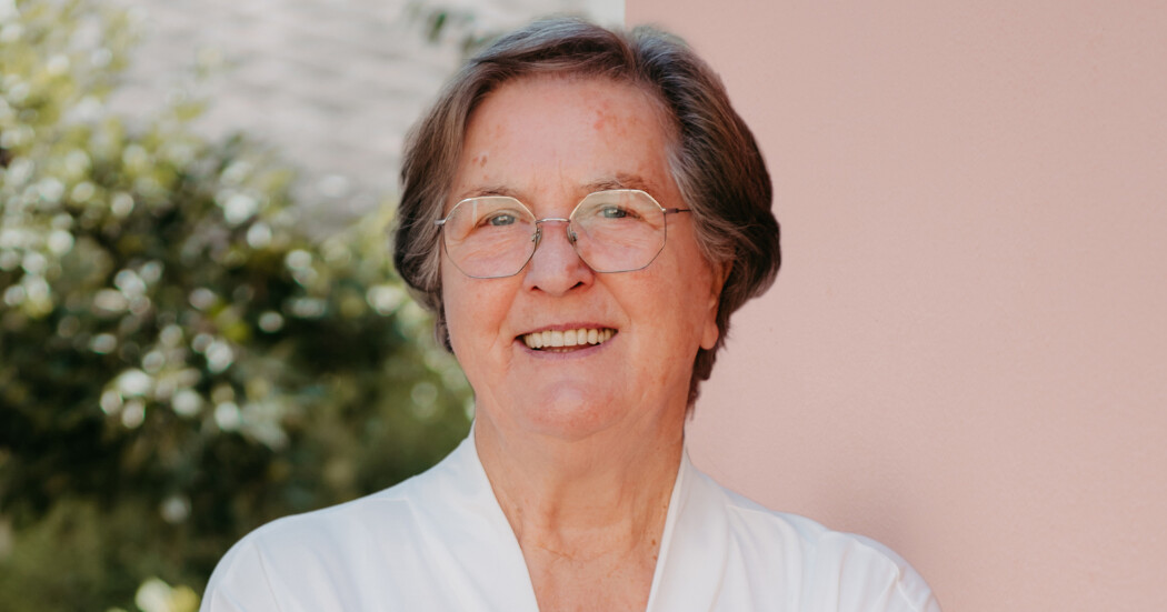 Person mit kurzen grauen Haaren, Brille und weißem Oberteil, lächelnd vor unscharfem Hintergrund aus Grünpflanzen und rosa Wand.