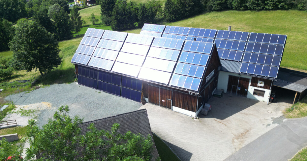 Holzgebäude mit großflächiger Solaranlage auf dem Dach, umgeben von grüner Landschaft, Bäumen und Kiesfläche im Hof.