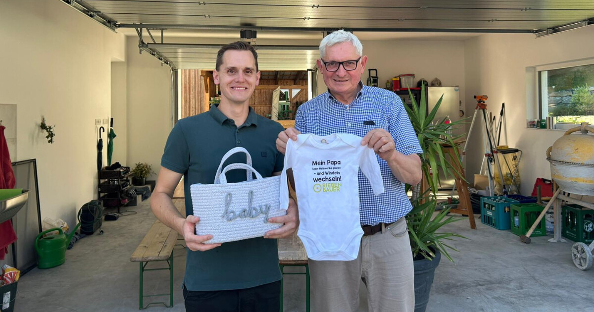 Zwei neutrale Personen in einer Werkstatthalle halten eine weiße 'baby'-Tasche und einen Baby-Body mit Spruch über Windeln wechseln.