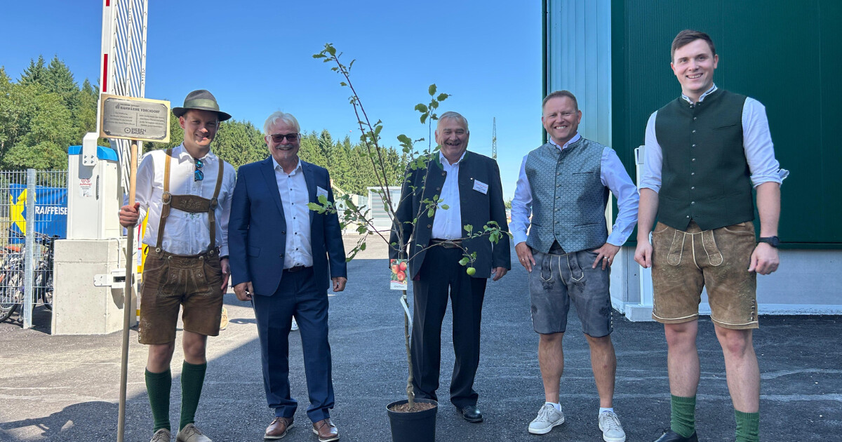 Fünf Personen stehen draußen in Trachten- und Businesskleidung um einen kleinen Baum im Topf.