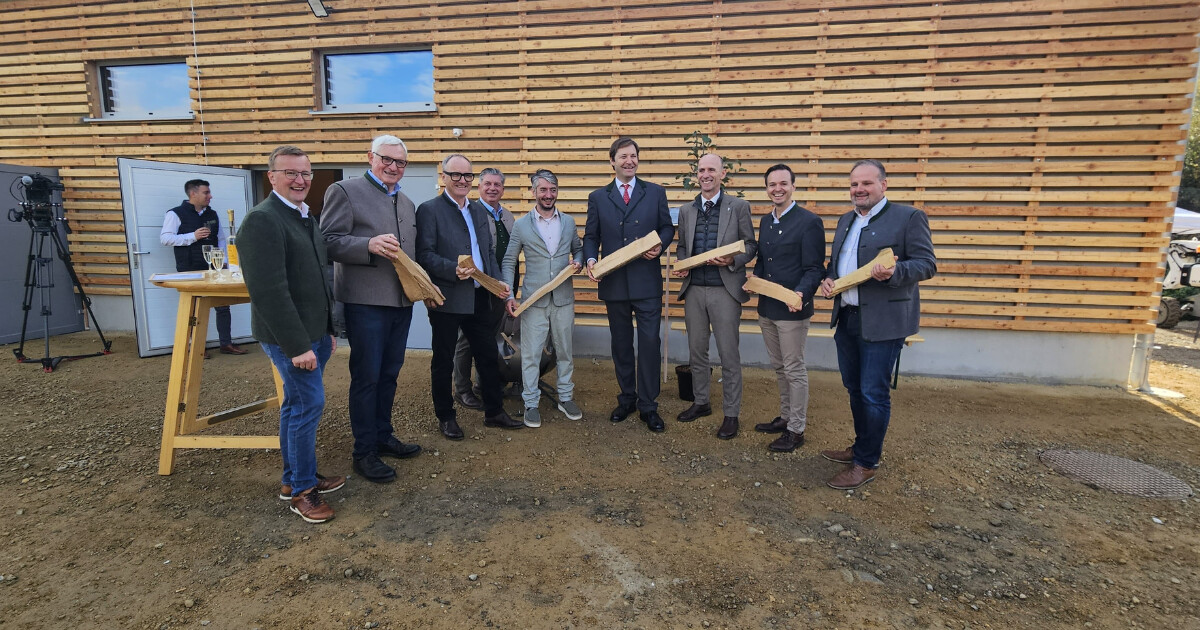 Gruppe von Personen in formeller Kleidung vor einer Holzfassade, hält lange Holzstücke als Zeremonie-Gegenstände.