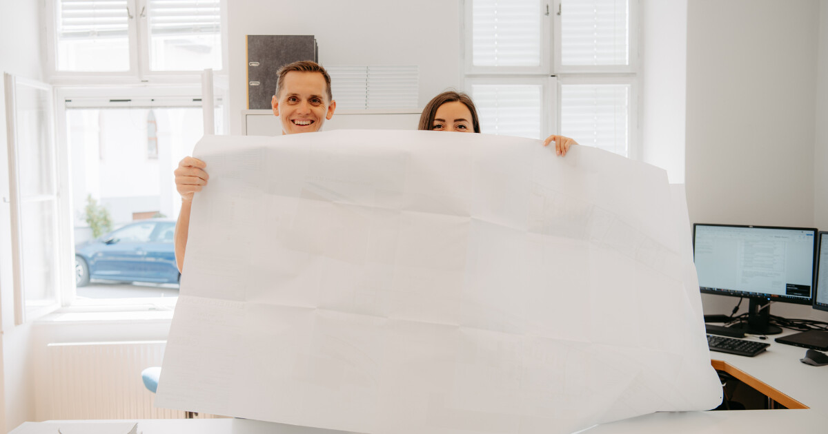 Zwei Personen halten eine große Bauzeichnung vor sich in einem hellen Büro mit Fenstern, Schreibtischen und Monitoren.