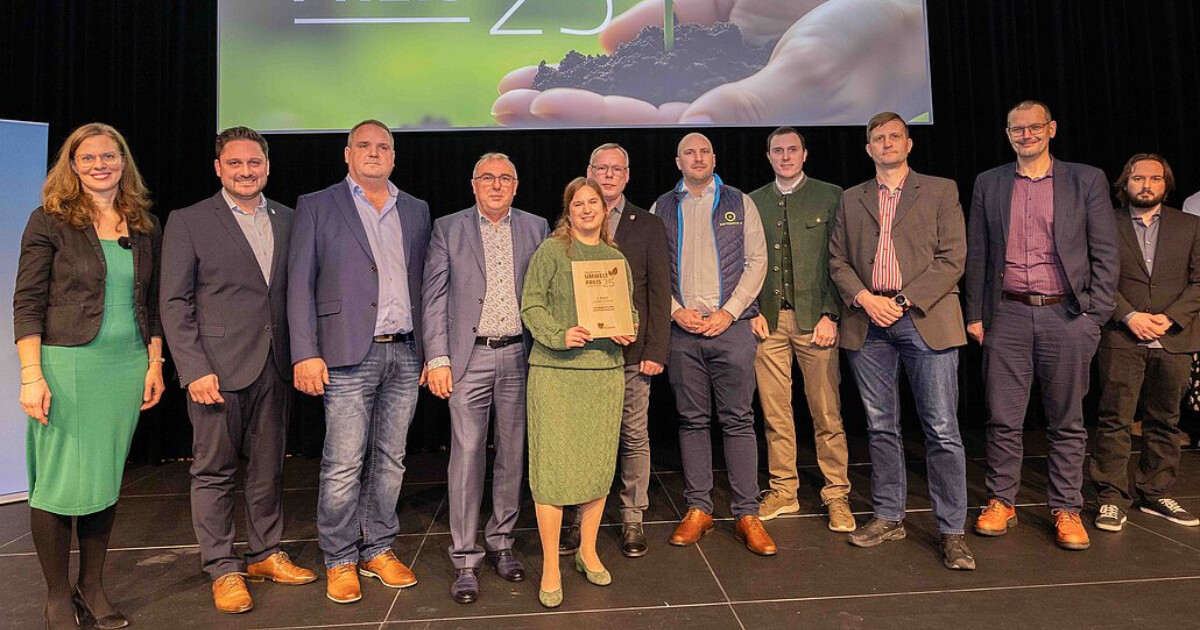 Gruppe von Personen steht auf einer Bühne; zentrale Person hält Urkunde bei Preisverleihung.