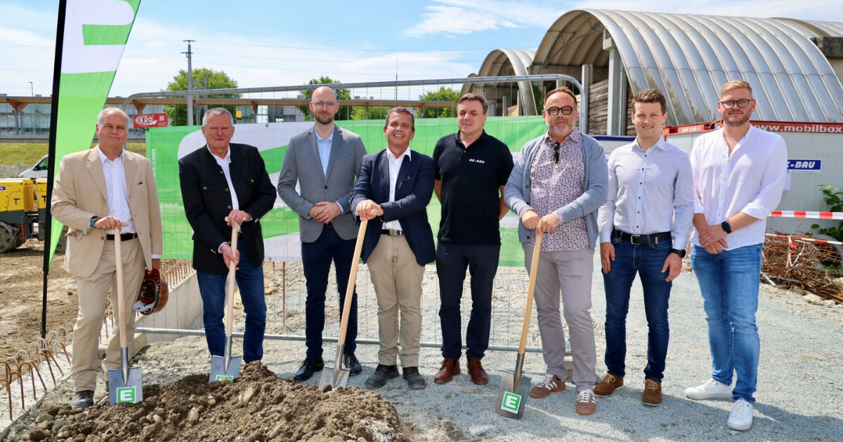 Acht Männer beim Spatenstich auf einer Baustelle; grünes Banner und halbrunde Bauhallen im Hintergrund.