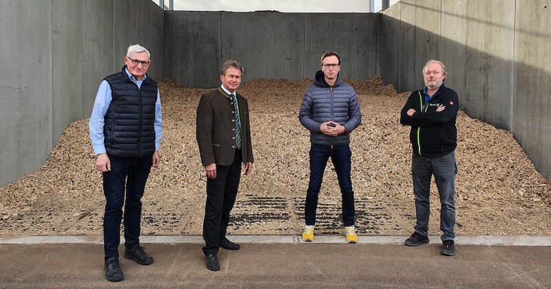 Vier Personen stehen in einer industriellen Umgebung vor einem Hackgutstapel, flankiert von Betonwänden.