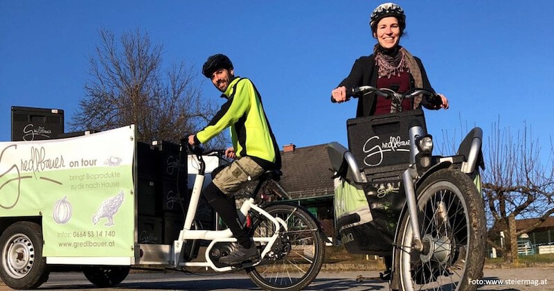 Zwei Personen mit Helmen fahren auf Fahrrädern; ein Lieferanhänger bewirbt Bio-Produkte, blauer Himmel im Hintergrund.