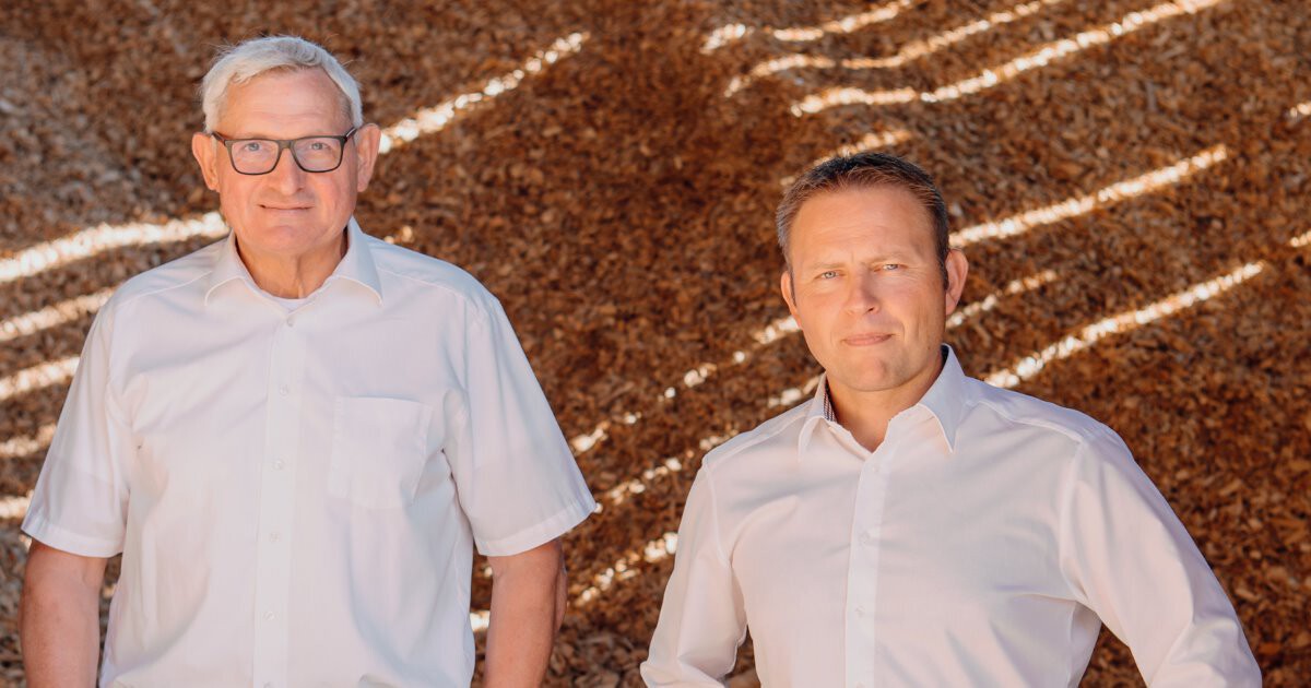 Zwei Personen in weißen Hemden stehen nebeneinander vor braunem Hintergrund aus Holzspänen und schauen in die Kamera.