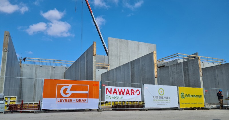 Baustelle mit Betonwänden, Baukran, Bannerwerbung und einer Person in Bauhelm; blauer Himmel im Hintergrund.