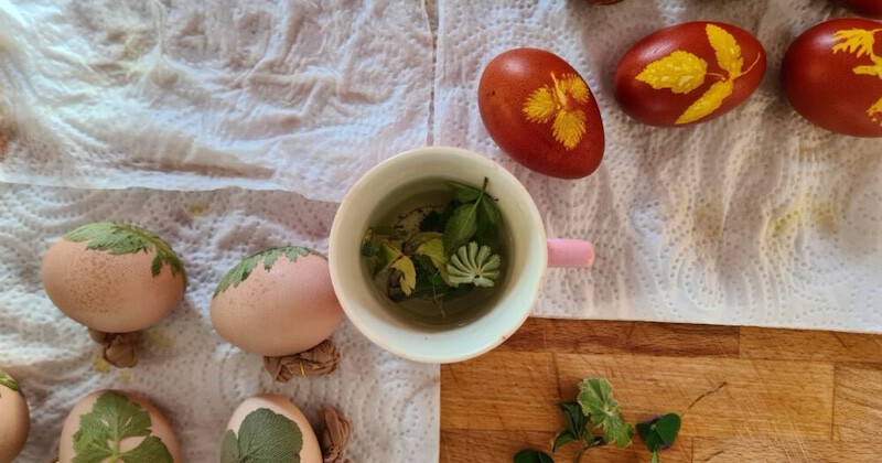 Ostereier in Braun und Rot mit Blättern verziert, daneben eine Tasse Kräutertee; Holzunterlage und Stofftuch mit Blattmustern.