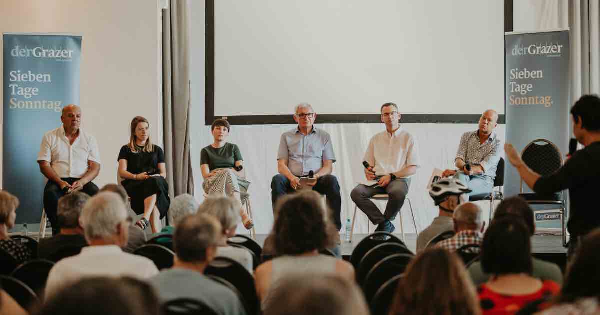 Sechs Panelteilnehmende sitzen auf einer Bühne bei einer Diskussionsrunde im Saal, vor Publikum; Banner links und rechts, Leinwand im Hintergrund.