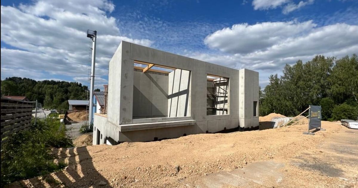 Rohbau aus Beton auf Kiesfläche; offener Baukörper mit großen Fenstern, Baugerüst, Baustellenmaterial und wolkigem Himmel.