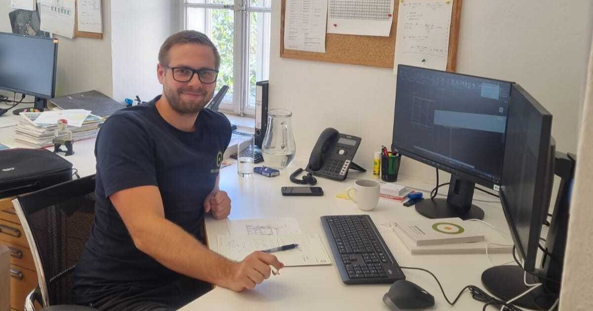 Person sitzt am Schreibtisch im Büro vor Computer und Monitor, mit Smartphone, Kaffeetasse, Notizpapier und Stiften.