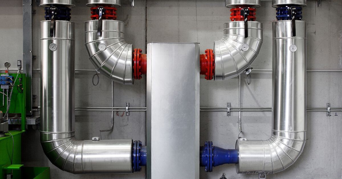 Industrielle Metallrohre und Verbindungen in einer Anlage, zentrales Stahlgehäuse, rote und blaue Flansche, Messgeräte an den Rohren.