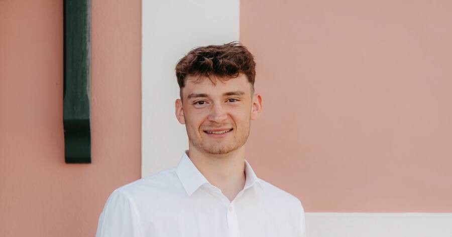 Person mit weißem Hemd lächelt vor einer pastellfarbigen Wand.