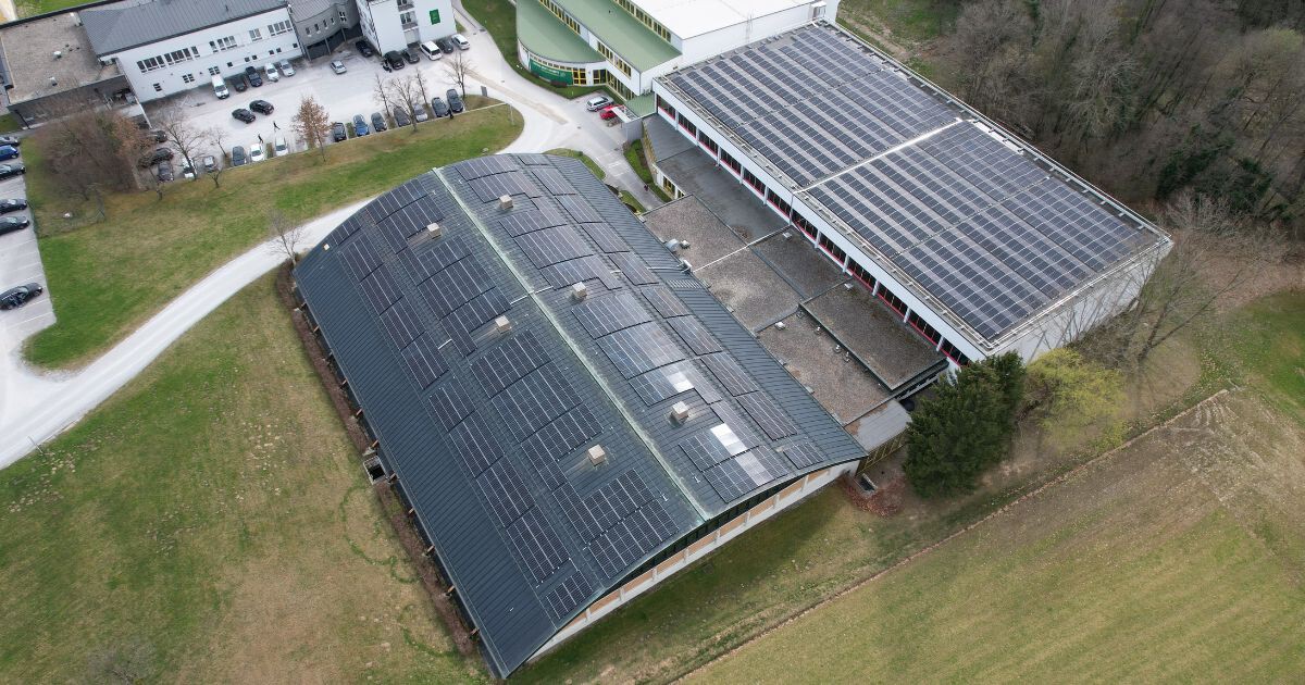 Drohnenaufnahme von Gebäudekomplexen mit Solarpanelen auf den Dächern, Parkplätzen und umliegenden Grünflächen.