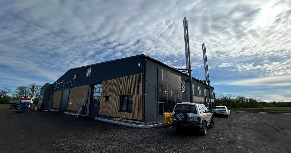 Modernes Industriegebäude mit Holzverkleidung und Solarzellen an der Fassade, zwei hohe Schornsteine, davor geparkte Fahrzeuge.