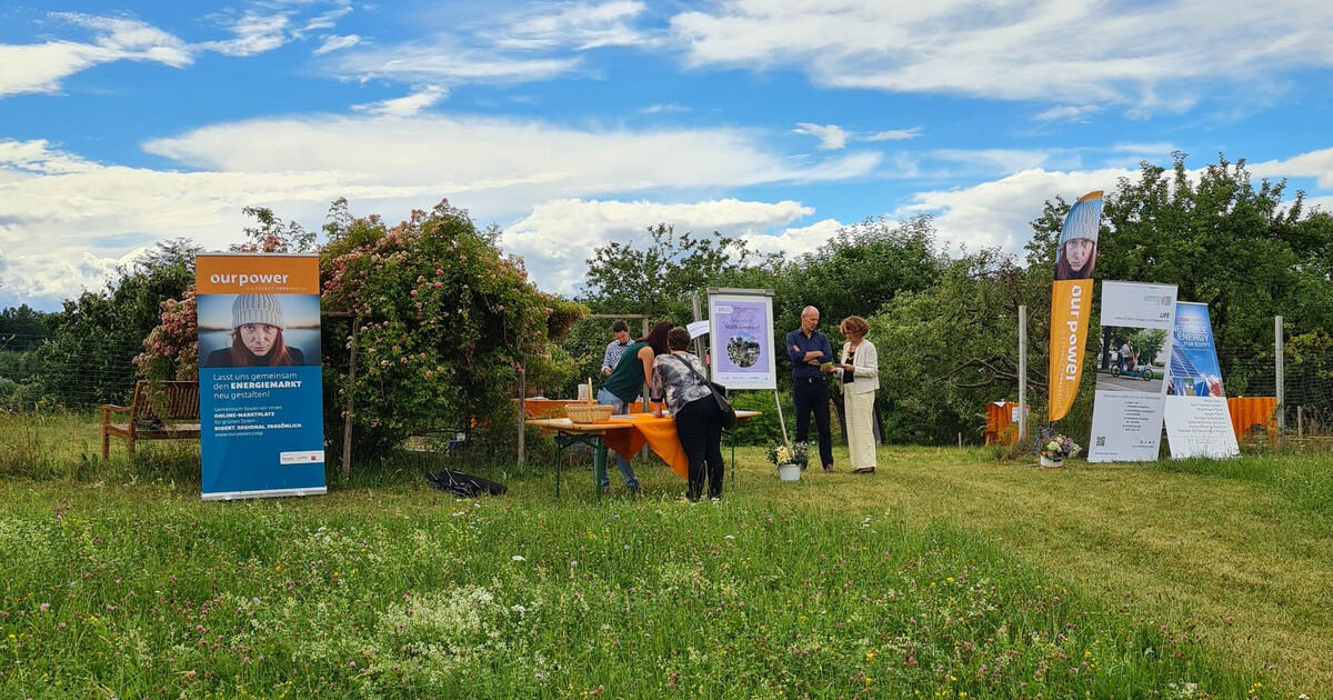 Außenveranstaltung auf grünem Gelände mit Infoständen, orangefarbenem Tischtuch und Bannerwerbung; mehrere Personen im Gespräch.