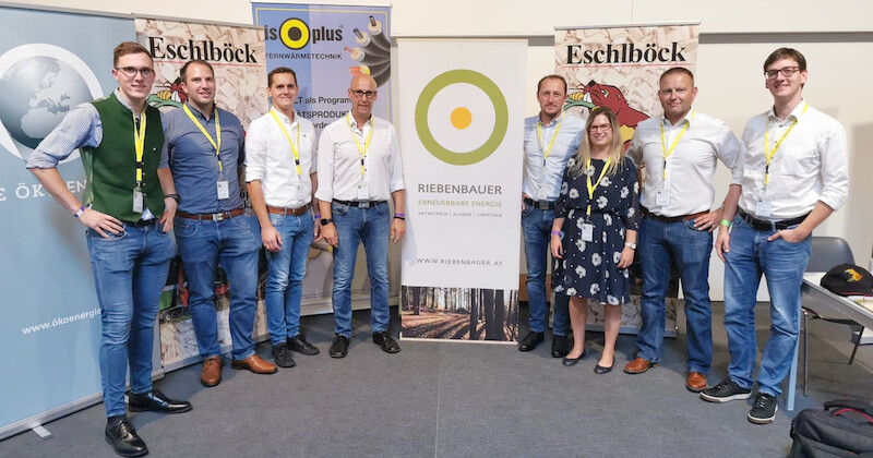 Neun Personen in Business-Casual vor Messestand mit Bannern; gelbe Lanyards, posieren für ein Foto.