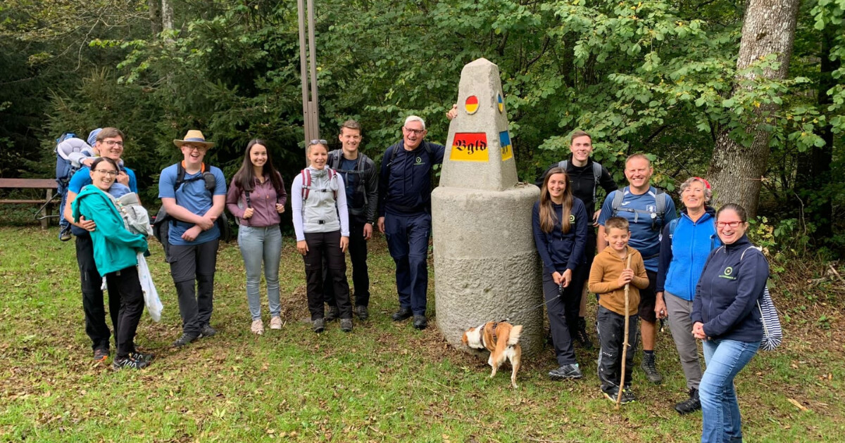 Gruppe von Personen vor einem Steinobelisk mit farbigen Aufklebern im Wald; Hund schnuppert am Boden.