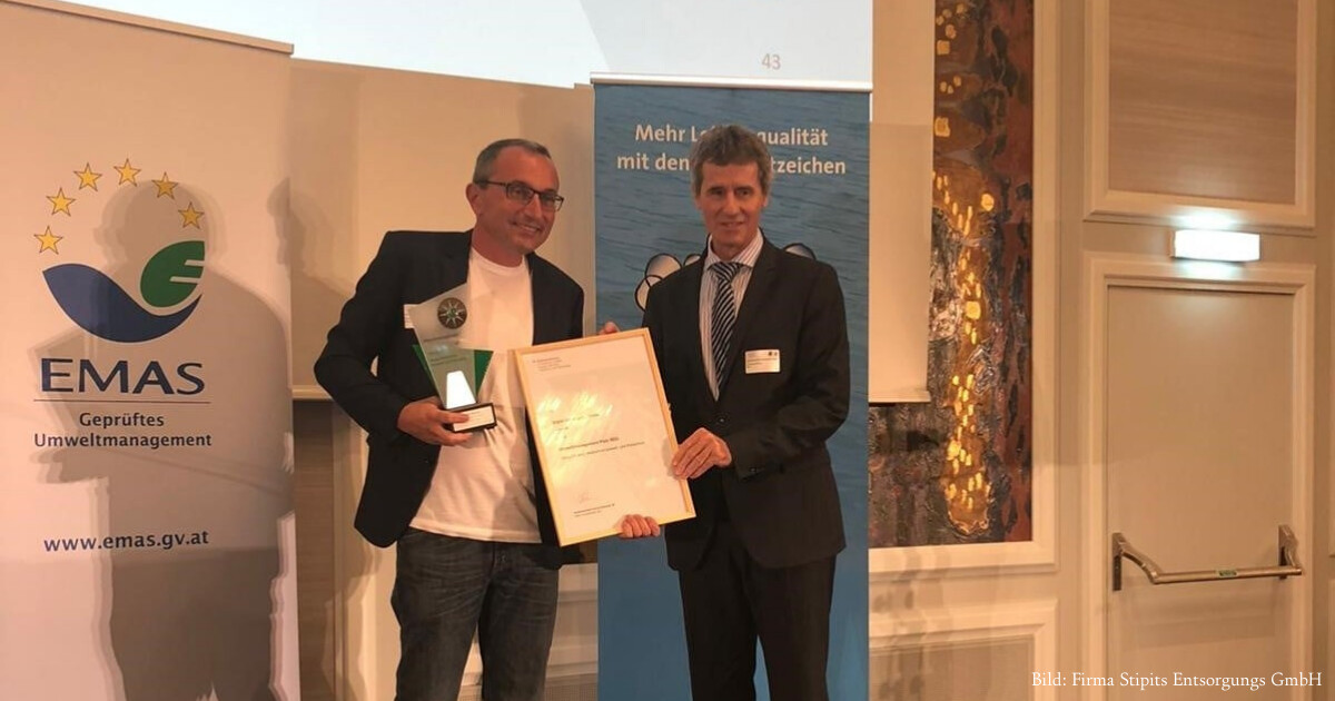 Zwei Personen in Anzügen erhalten Auszeichnung; eine hält Trophäe, die andere Urkunde – EMAS-geprüftes Umweltmanagement im Hintergrund.