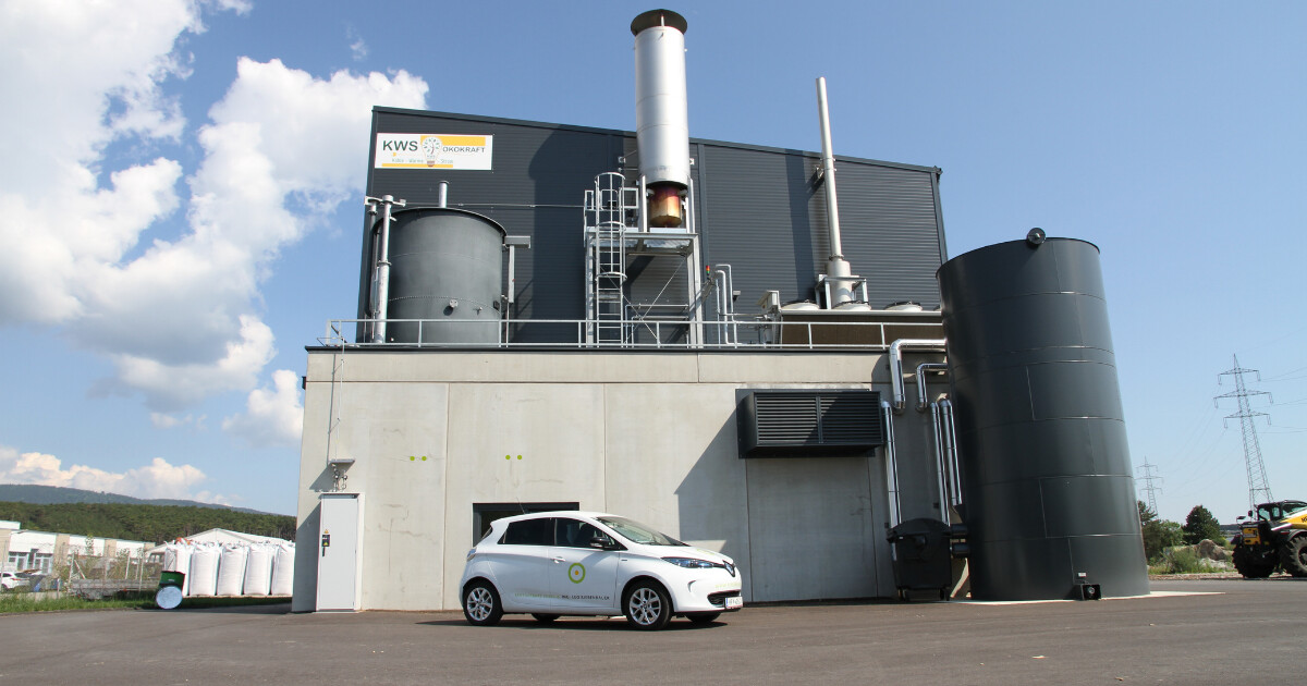 Industrieanlage mit Tanks, Rohren und Leitern; vorn ein weißes Auto mit Logo vor der Gebäudefassade.