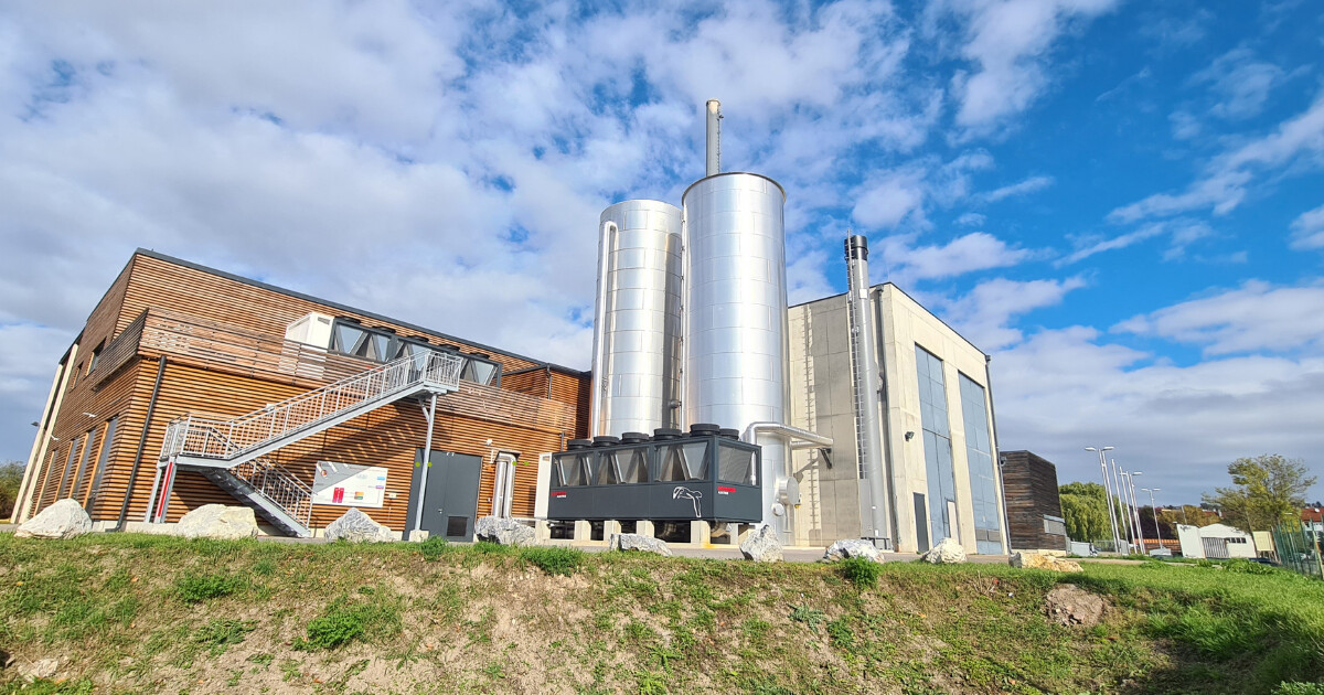 Industrieanlage mit Holzgebäude, zwei silbernen Pufferspeichern, technischer Container und Außenstiege; moderner Betonbau, grüne Wiese und blauer Himmel.