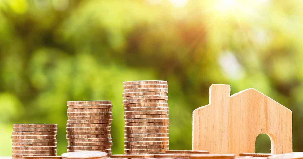 Stapel kupferner Münzen unterschiedlicher Höhe neben einem kleinen Holzhäuschen; unscharfer grüner Gartenhintergrund.