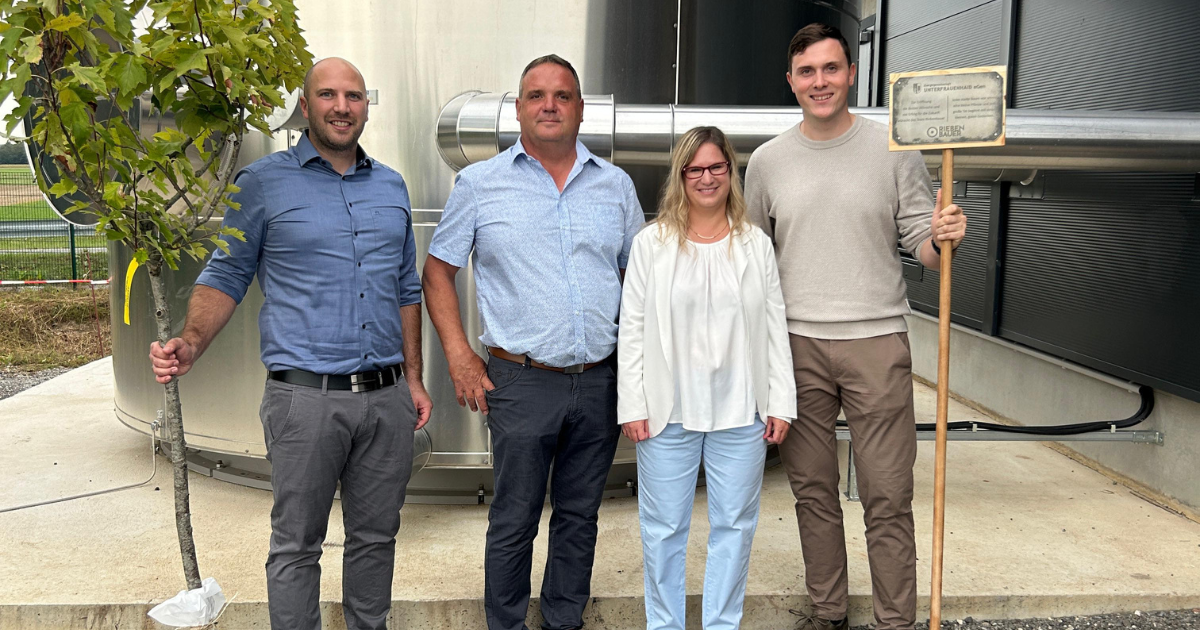 Vier Personen stehen vor einer industriellen Metallanlage; links hält eine Person einen Baum, rechts eine Person ein Schild.