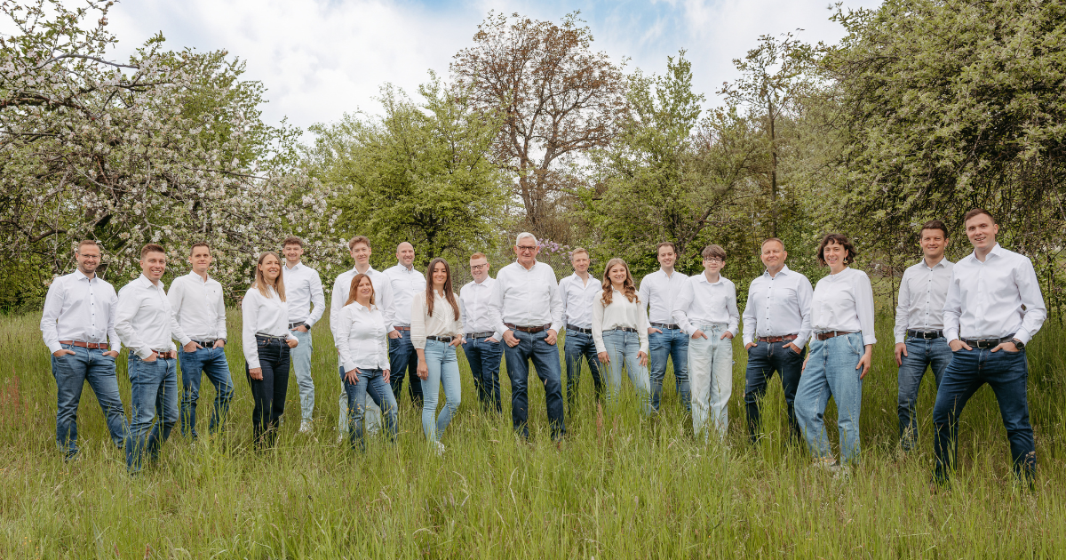 Gruppe von Personen in weißen Shirts und Jeans vor einem großen Stapel Baumstämme im Freien.
