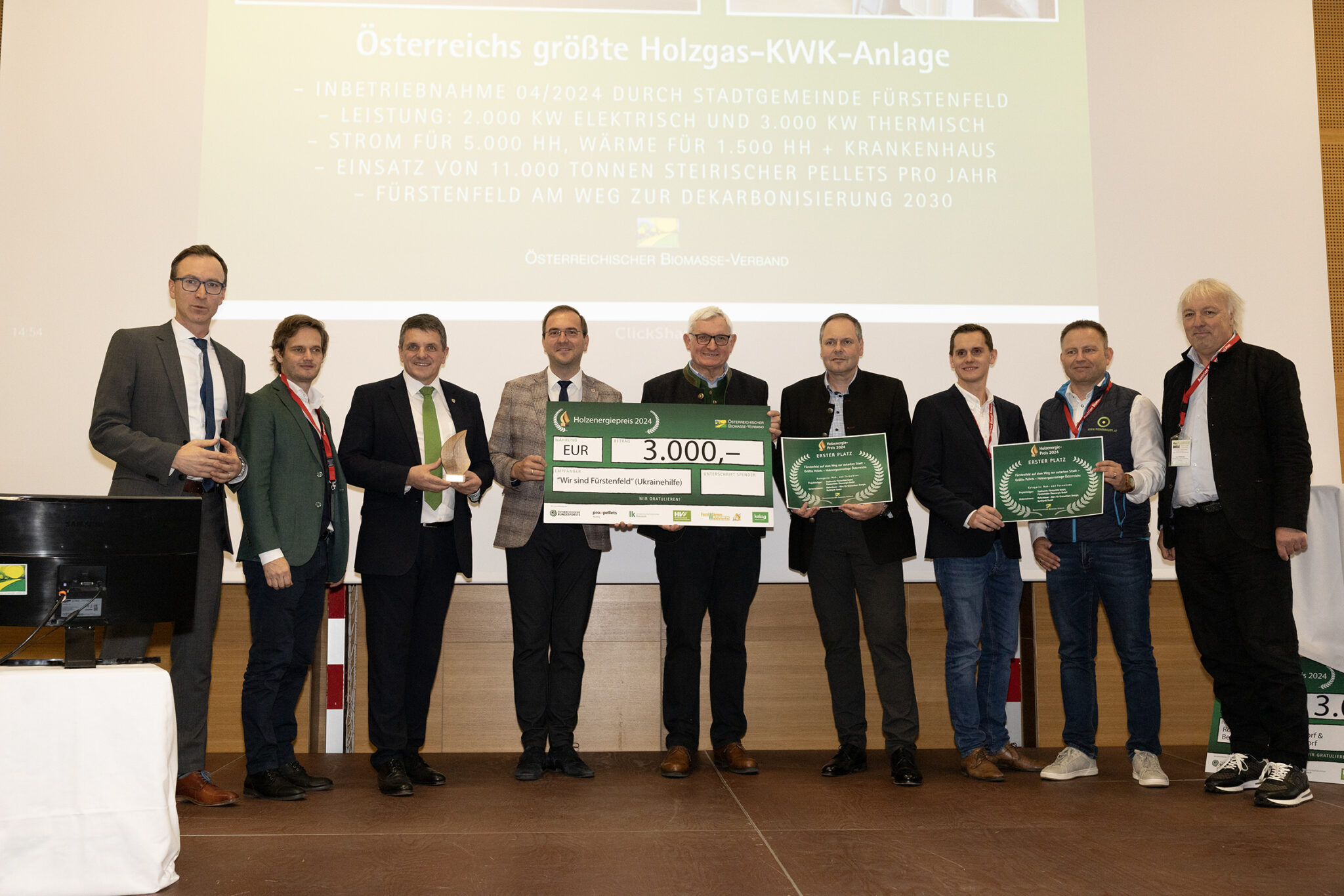 Neun Personen stehen auf einer Bühne bei einer Preisverleihung und präsentieren einen großen Scheck sowie Urkunden.