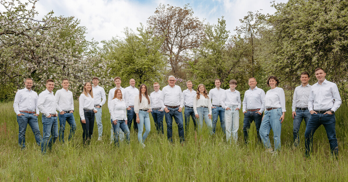 Gruppe von Personen in weißen Shirts und Jeans vor einem großen Stapel Baumstämme im Freien.
