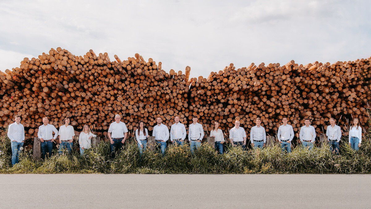 Gruppe von Personen in weißen Shirts und Jeans vor einem großen Stapel Baumstämme im Freien.