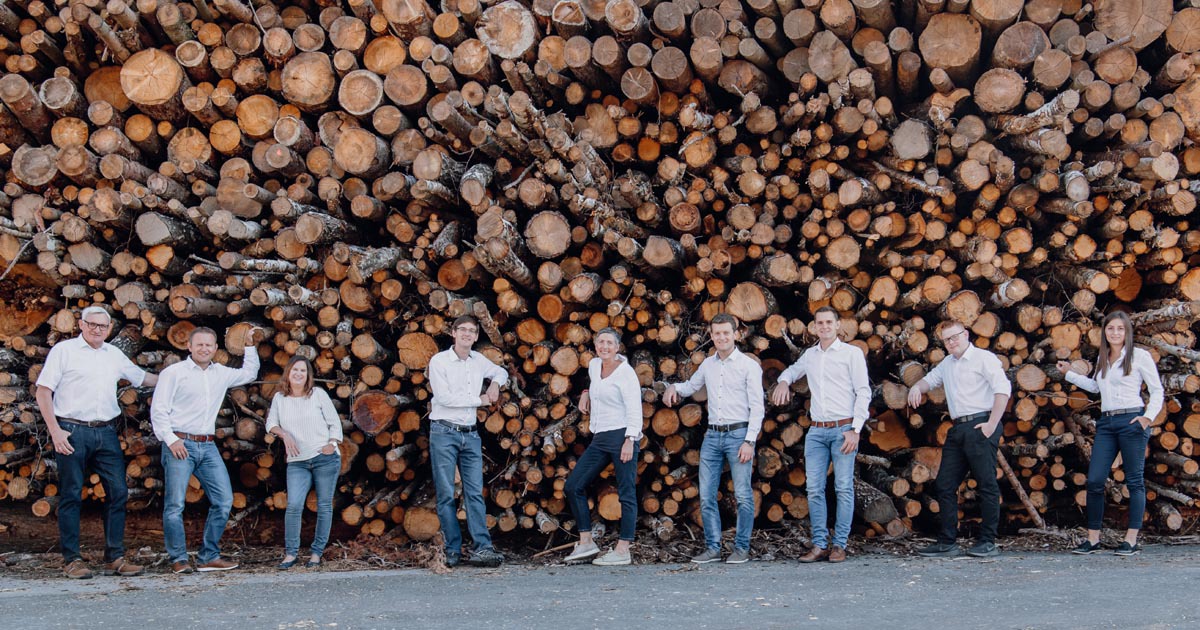 Gruppe von Personen in weißen Oberteilen vor einem großen Holzstapel; Outdoor-Teambild.